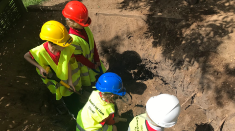 Four children, wearing hard hats and hi-vis bibs with 'Construction Worker' written on the back', dig a large hole at Elloughton Primary School  