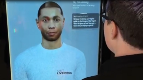A digital depiction of a young man wearing a white top with the Visit Liverpool logo on a large screen with some text captions in the top right corner. A man with glasses can be seen on the right using the service on screen.