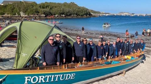 A group of people stand on a sandy beach beside a large blue wooden boat named 'FORTITUDE' with white lettering. A green tent is partially covering the boat. In the background, more people, boats, and water activities are visible in a bay or harbour, with vegetation and buildings along the distant shoreline.