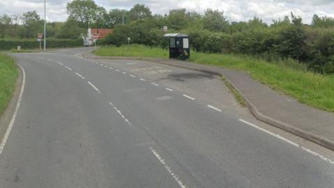 A rural road bordered by hedges with a bus stop and layby on the righthand side.