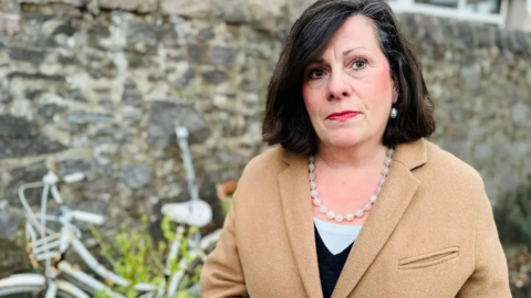 Councillor Mrs Jennifer Stewart stood in front of a wall and bike. She has a serious expression on her face, and is wearing a beige jacket and a necklace. 