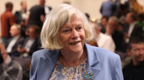 Ann Widdecombe smiles as she looks to the right. She is wearing a blue blazer and a flowery blue and white top.