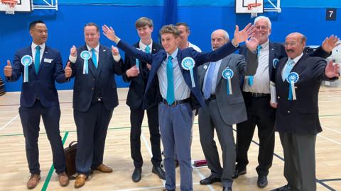 Reform UK candidates in the election count venue in Coalville, Leicestershire. They all sport blue rosettes and wear suits over white shirts and light blue ties.