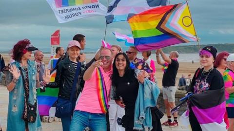 A group of people dressed in different colours and waving LGTBQ flags are standing by the beach in Weston-super-Mare. Several people are smiling towards the camera. 