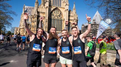 Four male runners in black sportswear waving to the camera in front of the Lincoln Cathedral. There are other runners in the background and a man in a hi-vis vest and a hat.