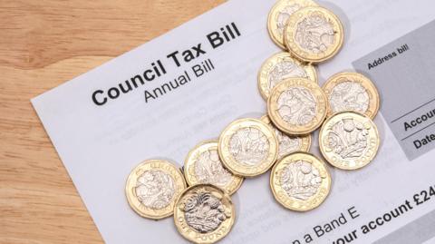 A council tax printed on white A4 paper lies in on a wooden table. This is a close up image and we see the heading 'Council Tax Bill' and the sub-heading 'Annual Bill'. We see some partial lines of more details. On top of the bill is a scattered pile of a dozen pound coins.