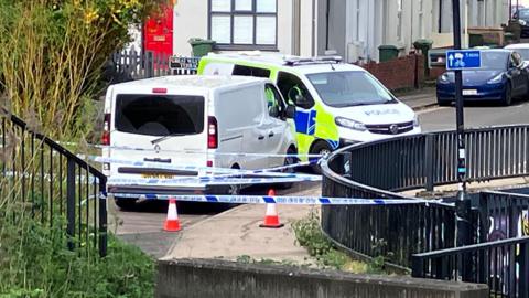 Two police vans are parked close to each other near a pavement with a dark fence running alongside it. There are cones and several lines of police tape running across the street.