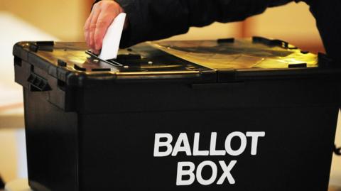 A hand slotting a voting paper into a black ballot box, marked in white lettering as 'Ballot Box'.