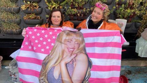 Scarlet and her mum Deborah sit on camping chairs in the queue. Scarlet has blonde hair which is in rollers. Deborah has long dark hair. They are both wearing orange survival ponchos over jumpers. They are holding a pink American flag with Sabrina Carpenter on it.