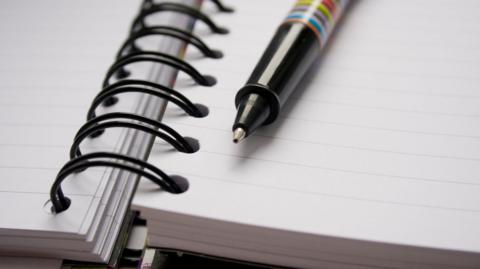 Close-up of an open spiral-bound notebook with lined pages. A diagonally placed pen featuring a colourful striped design rests on the notebook.