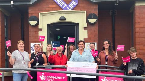 Members of the Streetlife charity smile and hold up National Lottery community fund pink flags and a cheque for £475,000 outside the entrance to Streetlife's base. 