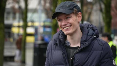 Jack Chadwick, who is wearing a black cap with a navy coat and black T-shirt, is smiling in this candid image in Piccadilly Gardens during filming. 