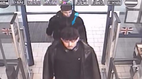 Two young men in black clothing are captured on CCTV looking forward while walking into a supermarket 