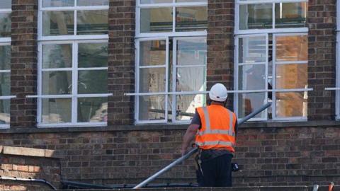 A man has his back turned to the camera, wearing a white hard hat and orange high-vis vest. He is holding a long metal pole and is standing at the foot of a school building. He is possibly standing on a flat roof.