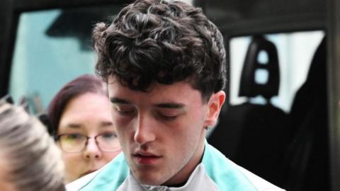 Layton Randle being taken into court. He has short brown curly hair and is wearing a white top with a green stripe and has a diamond earring. He is leaving a van and is being led by a security officer. 