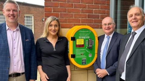 Four people standing next to a defibrillators mounted on a brick wall