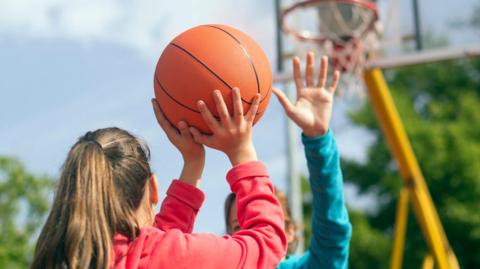 A young person seen from the back holding a basketball in the air as they get ready to take a shot. In the background there is a basketball hoops and another person standing in front of the first, holding their hand in the air.