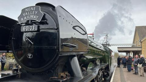 Crowds gather either side of The Flying Scotsman, as it shoots steam and smoke into the air. On the front of the locomotive a metal display plate reads "Nene Valley Railways".