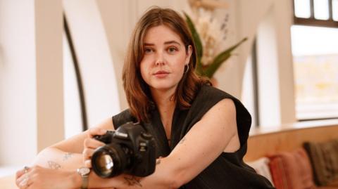 Artist Phoebe Wingrove sat with her camera. She is looking at the camera and wearing a short sleeved black top, with shoulder length brown hair. Behind her the room is decorated in neutral tones and there are large windows visible.