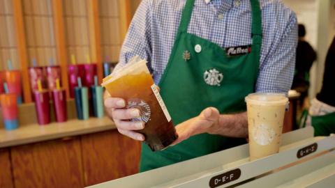 A barista places an iced coffee in the mobile pickup area at a Starbucks location in New York, US. The barista is dressed in a blue and white checkered shirt with a green apron with the Starbucks logo in the centre.