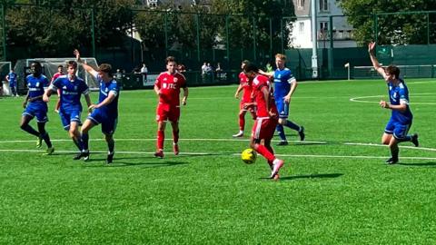Jersey Bulls players are dressed in a red kit with white stripes, and the Sheerwater players are in blue. A Jersey Bulls player has the ball and is getting ready to strike it goalwards.