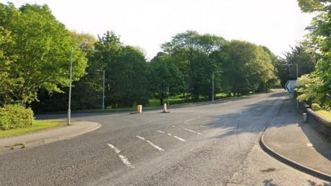 The junction between the B7076 and the A709. Trees surround the junction and there are pavements on the edges of the road. The road markings are faded and there are streetlights around the junction. 