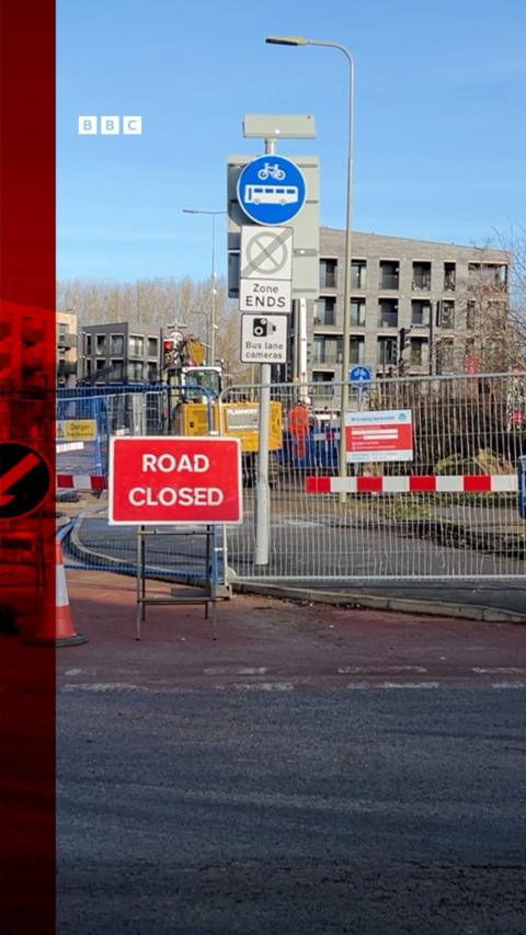 Road closed sign on a street corner with a buiding in the background.