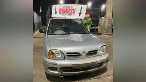 Matilda Walden in red beret and yellow high visibility jacket stands to the right of the silver Nissan Micra she sold for £400. She is in the event space at the Depot Mayfield in Manchester.