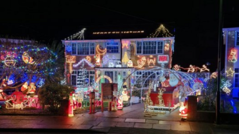Detached house covered in Christmas lights. There are snowmen and Father Christmases and reindeer and a sleigh with multicoloured rope lights and displays in the garden and on the house. There are the words Merry Christmas lit up on the roof of the house