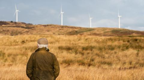 A man in a jacket and bunnet stares out across grassland towards four wind turbines in the distance