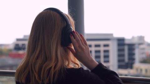 A woman look out over a cityscape while wearing headphones. She's facing away from the camera on the left side of frame. Her hand is touching the headphones as if immersed in music. She has blonde hair and pink nail varnish.