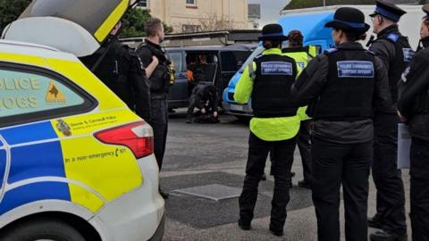 Seven police officers and community support officers in uniform are standing next to a parked police dog car in what looks like a car park. In the background there is a black van with an open door behind a man dressed in black bending down opening a rucksack.