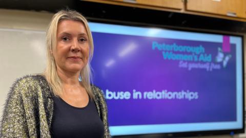 Mandy Geraghty stands in front of a screen showing a presentation from Peterborough Women's Aid that is headlined "Abuse in relationships". She has  blonde hair and wears a black tops under a black and yellow cardigan. She is looking straight at the camera.