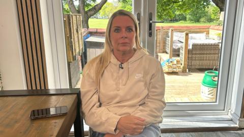 Lisa Barker sits in a living room in front of a patio door. She has shoulder-length blonde hair and is wearing a cream hoodie. Her mobile phone is on the table next to her. 