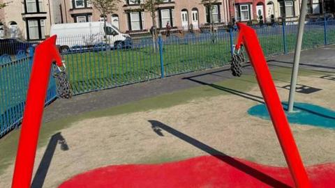 A children's playground in a park. A large red frame with two chains on the end is in the centre frame, it is empty, with no swing attached.