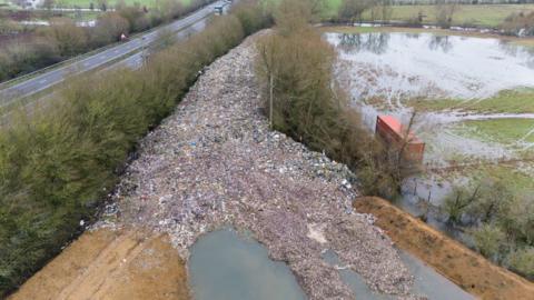 A large pile of waste next to a field and road. The waste stretches into the distance between two rows of trees.