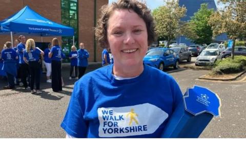 A woman smiles at the camera. She has short curly brown hair, and is wearing a blue t-shirt with the slogan 'We Walk for Yorkshire'