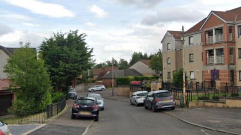 Scarrel Drive in Glasgow. Flats and houses like the streets. There are black railings between the gardens and the pavements and cars parked along the pavements.