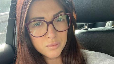 Charlotte Angel, who has long auburn hair and wears large red-framed glasses, poses for a selfie in the front seat of her stationary car.