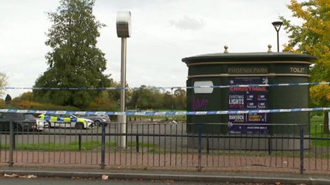 Police tape can be seen by a park. A police car is parked in the car park with other vehicles. A public toilet sits on the right of the image near a street light.