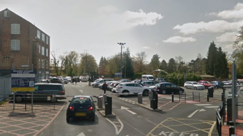 A dark car stopped at a car park barrier with its brake lights on. There are a number of cars parked in the car park.