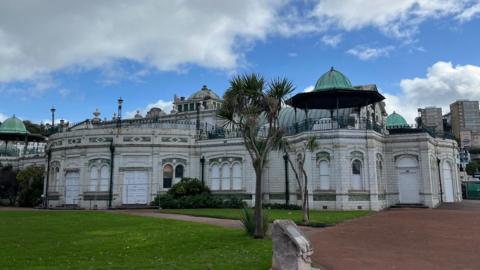 Exterior of Torquay Pavilion. The white building pictured on a sunny day. A palm tree and garden area with green grass can be seen in front of the white building.



