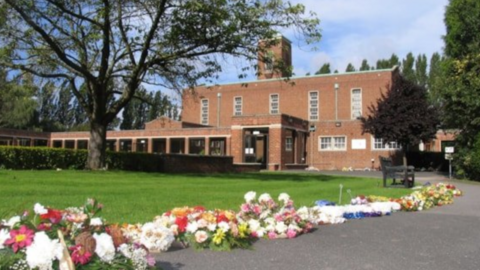 In the foreground there is a row of multi coloured flowers laying on the floor. they lead up to a brick crematorium building which has a flat roof and long rectangular windows. There is a large porch and a long outside corridor with windows. There are lots of tall trees and neatly cut grass. The sky is blue with some clouds.
