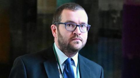 Oliver Mundell with short cut hair and a beard and glasses wears a navy suit with the blue shirt and a blue tie