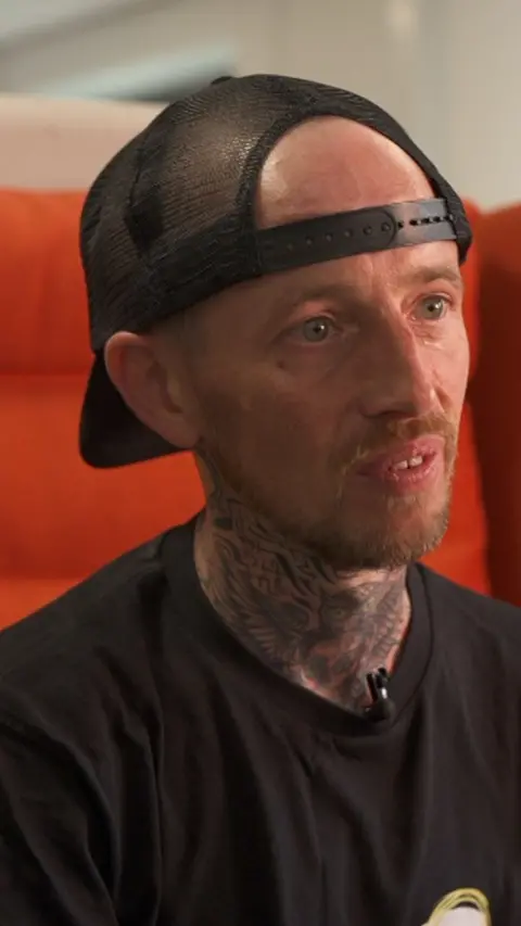Steven, a man in a black t shirt and black snapback cap sat on a large orange chair. He has a short red haired goatee.
