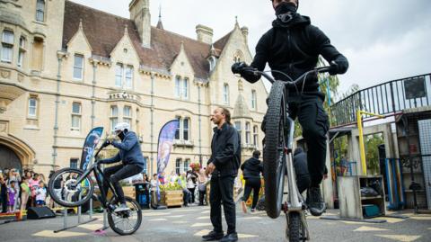 People dressed in dark clothing performing stunts on bikes with another person talking into a headset microphone with spectators in the background