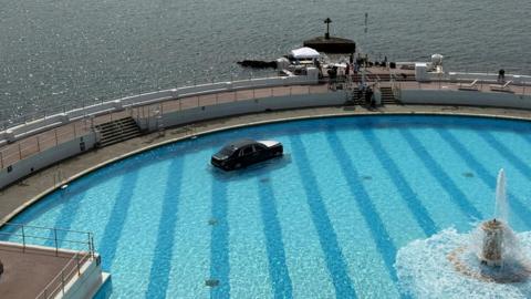 A Rolls Royce car on a submerged plinth in Tinside Lido in Plymouth on a sunny day. A fountain in the middle of the pool is spouting out water. The car is mostly black with a silver bonnet. A film crew is gathered on the outside edge of the pool.
