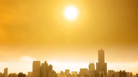 A panoramic view of a city with the sun directly overhead, casting an orange glow across the sky from the intense heat