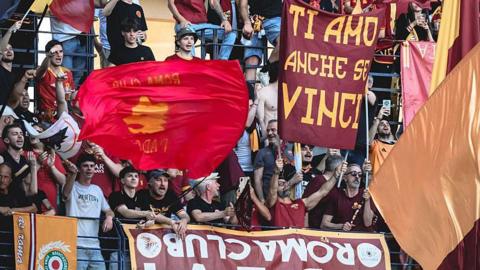 Roma fans in red club shirts, holding up banners and flags in a stadium