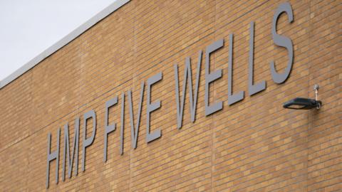 A sign for HMP Five Wells displayed on a large brick wall.